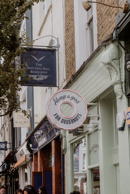 Close-up view of the exterior of a commercial building on a busy street during daytime, showing two illuminated signs hanging above the sidewalk. The first sign reads 'The Notting Hill Bookshop' with a dark blue background and white text, attached to a metal bracket extending from the wall. The second sign, designed as a large, round doughnut with pink icing and hexagonal hole in the center, displays the message 'Always a good for doughnuts' in black lettering. Surrounding the signs are brick and light-colored wooden building facades, with a window partially visible on the upper floor. The street appears lively with pedestrians walking nearby, and a canopy extending over the sidewalk. The scene captures the essence of a quaint, characterful area suitable for home relocation or moving services in Notting Hill, with an emphasis on urban storefronts and pedestrian environment.