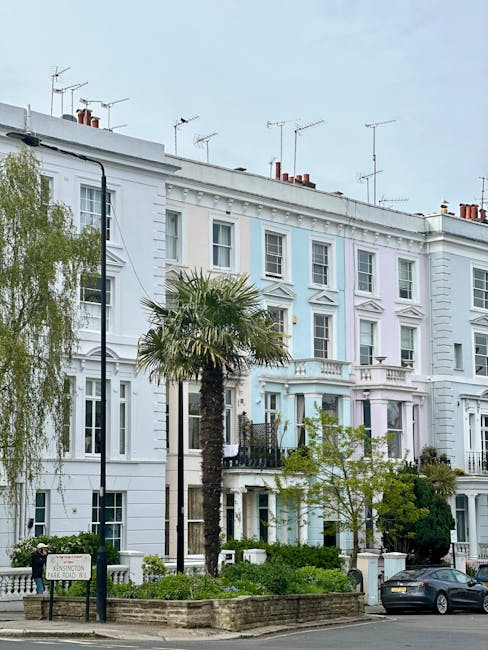A row of white, Victorian-style terraced houses with detailed architecture, including large windows and decorative cornices, is seen in a residential area. Several chimneys and TV antennas are visible on the rooftops. In the foreground, a small garden with a tall palm tree, other greenery, and low brick walls separates the pavement from the properties. A black street lamp stands near the garden, and a parked dark grey car is on the street. The image depicts a daytime scene with natural lighting, highlighting the exterior of the homes. The setting is characteristic of a London neighbourhood, suitable for house removals and home relocation services offered by Man and Van Notting Hill, as seen in the context of packing and moving logistics within a typical urban environment.