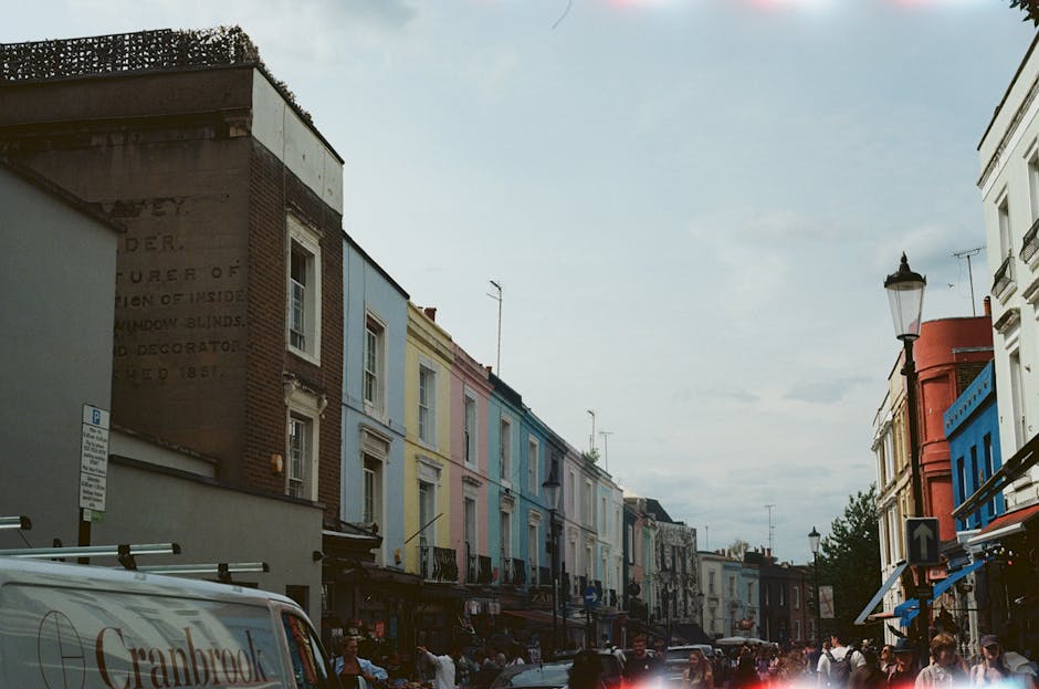 Exterior view of a row of Victorian-style terraced houses with decorative architectural details and multiple windows, painted in pastel blue and vibrant red colors. The buildings are fitted with tradi