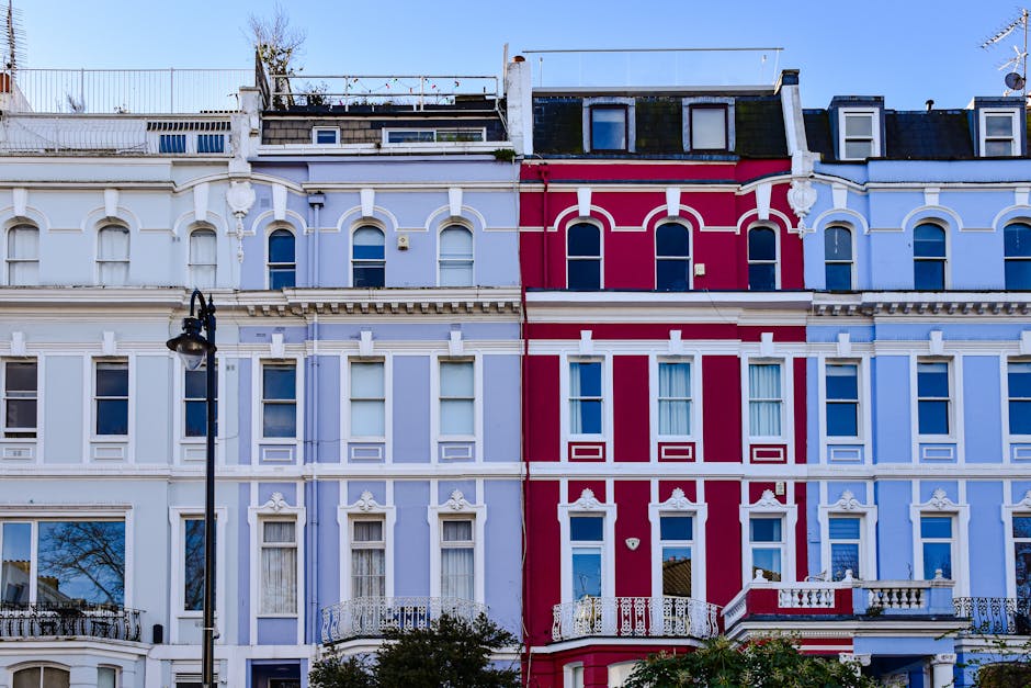 The image shows a close-up view of a row of multi-storey residential buildings with ornate façades, featuring white and red decorative details, situated in Notting Hill. In the foreground, a black street lamp is visible, positioned on the pavement in front of the buildings. The top floors of the buildings include white window frames, some with small balconies and railings, and the roofs are partially visible with dormer windows and rooftop access structures. The building facades showcase intricate molding and trim, emphasizing their Victorian architectural style. The scene is illuminated by natural daylight, highlighting the vibrant colours and architectural details of the houses, which are likely part of a relocation or home moving process supported by a professional removals service, such as Man and Van Notting Hill, specialising in house removals and furniture transport services.