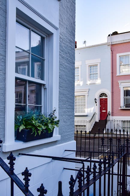 A close-up view of a white-painted exterior wall of a terraced house, featuring a white-framed sash window with a flower box filled with green plants and foliage on the windowsill. In the foreground, a black wrought iron railing with decorative finials encloses a small front garden area, which is part of a street lined with similar terraced houses. The image captures the window and railing from a slightly elevated perspective, with other houses visible across the street, including a white house with a black door and a pink house with white trim. The scene is illuminated by natural daylight, emphasizing the tidy and well-maintained appearance of the residential buildings, representing a typical urban setting suitable for home relocation or furniture transport activities undertaken by professional moving services such as Man and Van Notting Hill, especially when planning packing and loading processes for house removals.