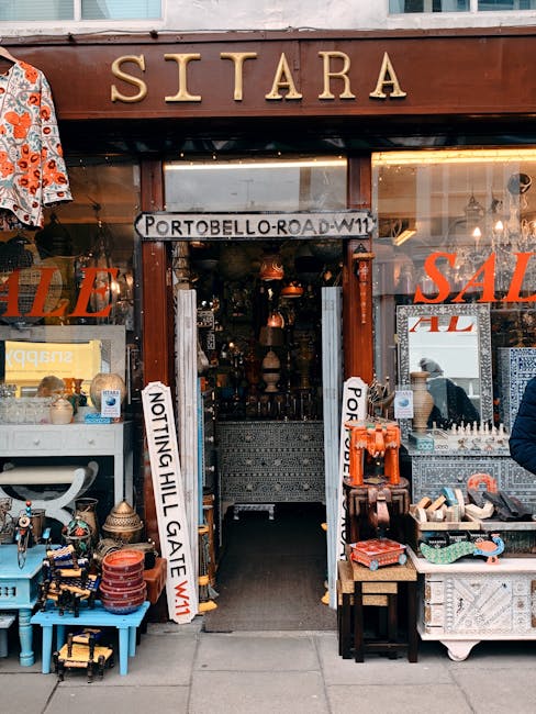 The entrance to a shop named Sitara is visible with a wooden signboard displaying the name in large gold letters. The shop is located on Portobello Road in Notting Hill, as indicated by a street sign above the doorway. The shopfront has large glass windows showcasing various items, including decorative ceramics, textiles, and jewelry, with sale signs partially visible. Inside, the interior features warm lighting and densely packed shelves, creating a cozy shop environment. Outside, on either side of the entrance, are display tables and racks filled with small decorative objects, pottery, and accessories. This retail setting is part of a busy street scene, with pedestrians walking nearby. The image, while primarily depicting a retail storefront, subtly connects to home relocation themes through the presence of various household and decorative items, which could be relevant to packing and moving services. Man and Van Notting Hill offers removals and furniture transport in this area, assisting clients with packing and home relocation logistics.