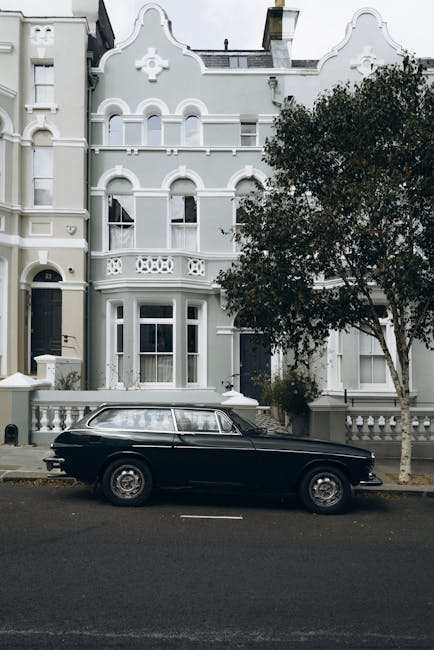 A close-up view of a white-painted exterior wall of a terraced house, featuring a white-framed sash window with a flower box filled with green plants and foliage on the windowsill. In the foreground, a black wrought iron railing with decorative finials encloses a small front garden area, which is part of a street lined with similar terraced houses. The image captures the window and railing from a slightly elevated perspective, with other houses visible across the street, including a white house with a black door and a pink house with white trim. The scene is illuminated by natural daylight, emphasizing the tidy and well-maintained appearance of the residential buildings, representing a typical urban setting suitable for home relocation or furniture transport activities undertaken by professional moving services such as Man and Van Notting Hill, especially when planning packing and loading processes for house removals.