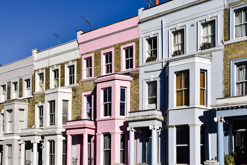 A row of traditional terraced houses in Notting Hill with ornate bay windows and decorative moldings, painted in soft pastel colors including white, light blue, and pale pink. The buildings have brick facades with architectural details, and some windows feature flower boxes. Bright sunlight casts shadows on the façades, illuminating the residential street. Nearby, a moving company like Man and Van Notting Hill may be involved in home relocation or furniture transport, with the process of packing and loading visible in the context of house removals. The scene highlights the typical urban environment where household items, including boxes, furniture, and packing materials, are temporarily stored outside or being moved between properties or onto a van for transport, demonstrating the logistical aspects of domestic moving services in urban settings.