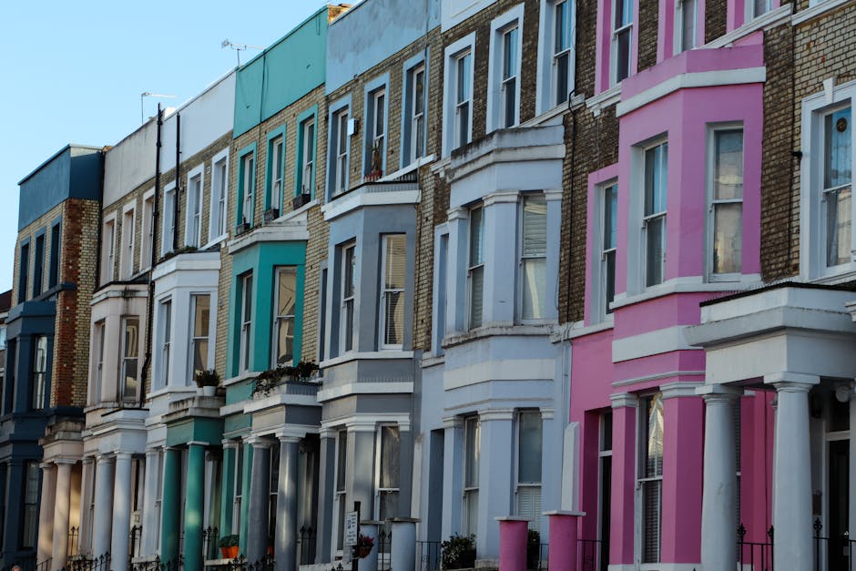 A row of traditional terraced houses in Notting Hill with ornate bay windows and decorative moldings, painted in soft pastel colors including white, light blue, and pale pink. The buildings have brick facades with architectural details, and some windows feature flower boxes. Bright sunlight casts shadows on the façades, illuminating the residential street. Nearby, a moving company like Man and Van Notting Hill may be involved in home relocation or furniture transport, with the process of packing and loading visible in the context of house removals. The scene highlights the typical urban environment where household items, including boxes, furniture, and packing materials, are temporarily stored outside or being moved between properties or onto a van for transport, demonstrating the logistical aspects of domestic moving services in urban settings.