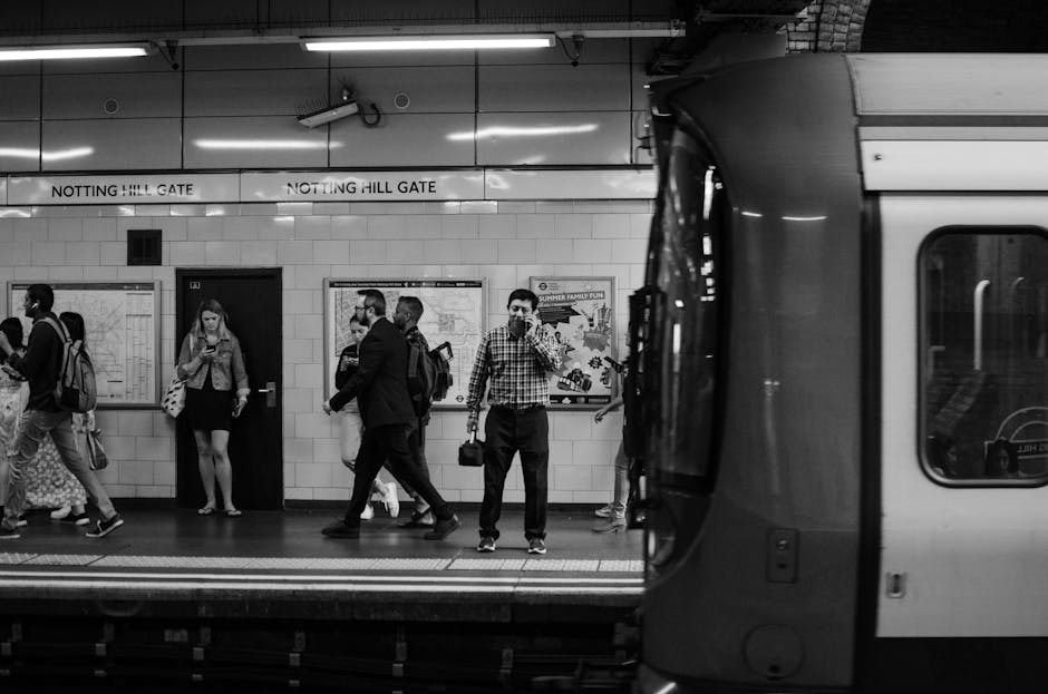 Black and white photograph of Notting Hill Gate underground station platform with several passengers waiting for the train, some carrying backpacks and shoulder bags, while others stand near maps and signage on the tiled wall. A man in a checked shirt and trousers stands in the center, holding a bag and waving at the camera, with a friendly expression. The platform features signs reading 'Notting Hill Gate' above the tiled wall, and the edge of the train platform is visible in the foreground with the train partially seen on the right side of the image. This scene captures the typical busy atmosphere of a London tube station, which could be associated with home relocation or moving logistics involving passenger transit and transport connections, relevant to services like those provided by Man and Van Notting Hill.