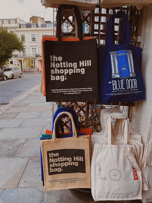 A display of reusable shopping bags hanging on a metal grid outside, with a street and residential buildings visible in the background. The bags are in various colors and materials, including black, blue, beige, and off-white, some featuring printed logos or text such as 'the Notting Hill shopping bag' and 'BLUE DOOR.' The bags are stacked and draped over the grid, with some positioned at different angles. The scene is outdoors, with natural daylight illuminating the bags, which are associated with local shopping or home relocation activities, and could be used in moving or packing processes supported by a professional removals service like Man and Van Notting Hill.