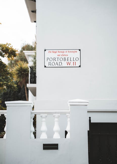 A white exterior wall of a residential property with a black metal mailbox mounted on the wall near the entrance. The wall features a decorative white balustrade on top of a low white fence with classical-style pillars. Above the fence, part of a door or window frame is visible on the left side of the image. A street sign is attached to the wall, indicating the location as Portobello Road in Notting Hill, marked with the Royal Borough of Kensington and Chelsea. The sign is white with black and red text. The environment includes some greenery with trees and bushes visible in the background, indicating an outdoor setting on a residential street. The lighting suggests daylight, and the scene captures a typical step in a home relocation process, where careful packing, loading furniture, and moving equipment may be involved by professional removals services such as Man and Van Notting Hill.