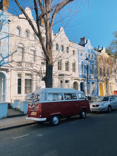 A vintage red and white Volkswagen van is parked on the street in front of a row of colorful Victorian-style terraced houses in Notting Hill. The van has a bike rack mounted on its rear and is positioned on the pavement, partially blocking the curb. To the right of the van, there are modern cars parked along the street. The houses behind feature ornate architectural details, with facades painted in pastel shades such as blue, yellow, and white, and have large sash windows with decorative trim. A leafless tree stands directly in front of the houses, with its branches extending over the van and street scene, indicating a winter or early spring season. The lighting suggests a clear, sunny day, providing natural illumination to the scene. This setting aligns with typical home relocation activities, where Man and Van Notting Hill might assist with furniture transport and packing during house removals in the area. The image captures a typical moving environment outside residential properties, showcasing the urban street and logistical aspects of home relocation.