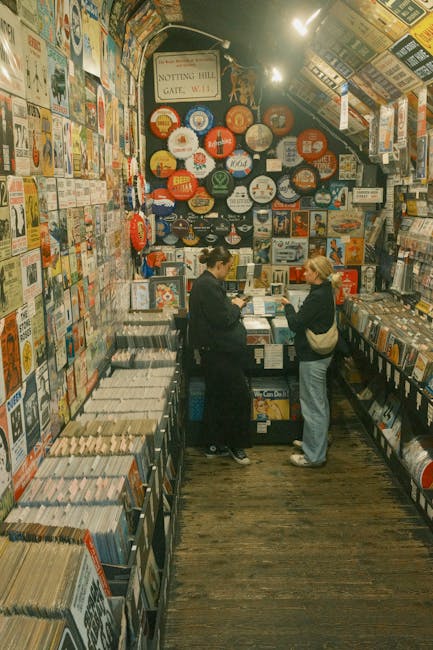 Inside a small, densely stocked shop with walls covered in colorful posters, signs, and illustrated advertisements, two women are engaged in conversation near a counter filled with books and magazines