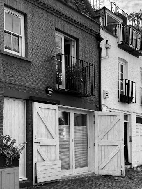A black and white photograph of a residential building facade showing two floors with balconies. The ground floor has a large glass door with horizontal blinds, flanked by wooden shutters that are partially open, revealing the interior space. A small wooden bench is positioned in front of the door, and a potted plant is placed to the left near the window. The first floor features two French doors with small balconies equipped with metal railings, and some plants are visible on the balconies. The building material includes brick and painted surfaces, with some areas of the facade showing texture and weathering. The scene captures a typical urban residential setting, relevant to house removals and moving services, illustrating the process of home relocation and furniture transport from a building with stairs, parking challenges, or lift limitations, as featured on the page 'Stairs, Parking or Lift Problems? Notting Hill Solutions' by Man and Van Notting Hill.