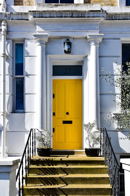 A white residential building features a bright yellow front door accessed by four stone steps with black metal railings on either side. The door is framed by decorative white columns and a rectangular overhead light fixture. Potted plants with small shrubs are placed on each side of the entrance on the steps. Above the door, the house number '27' is displayed on a dark plaque. To the left of the door, there is a window with a white frame, and the exterior walls are decorated with classical architectural details. The overall setting is well-lit, with sunlight casting shadows on the steps, suggesting a clear day. The scene depicts typical elements involved in house relocation or moving logistics, with the focus on the property's entrance and entryway preparations.
