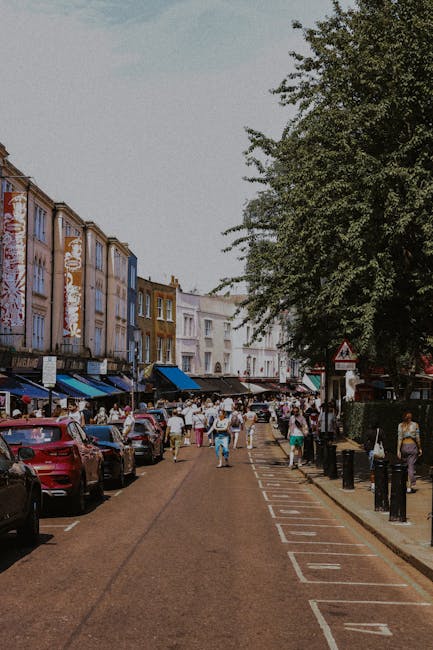 A street scene in Notting Hill during daylight hours, showing a busy pedestrian area with numerous people walking along the pavement and crossing the road. On the left side, a row of parked cars lines the street, with some partially visible behind pedestrians. The buildings on the left feature decorative facades, with several retail shops or cafes at ground level, identifiable by awnings and signage, and multi-storey residential or commercial structures above. To the right, a large leafy tree provides shade over the sidewalk, and a series of black bollards separates the pedestrian zone from the roadway. The road surface appears to be gravel or cobblestone, with marked parking bays and a narrow loading zone visible. The scene captures typical urban activity related to home relocation or furniture transport, as it may be part of a moving process through narrow streets common in Notting Hill, with the potential involvement of professional removals services like Man and Van Notting Hill.
