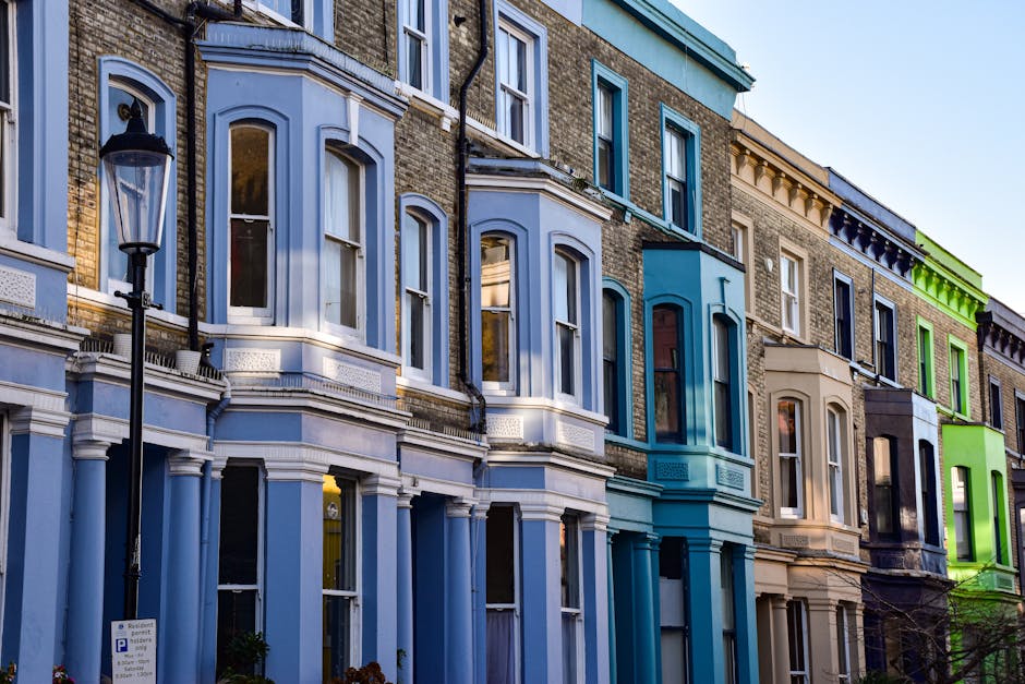 A row of multi-storey Victorian-style terraced houses with bay windows and decorative architectural details, featuring facades painted in various pastel shades including light blue, beige, and vibrant green. A black wrought iron street lamp stands on the pavement in front of the buildings, which are set beside a narrow street with a parking sign visible at the curb. The scene is captured during daylight with clear skies, highlighting the color contrasts and the window reflections. The image illustrates the urban residential environment typical of Notting Hill, and may relate to home relocations or furniture transport, with the buildings ready for a move when hosting packing and loading processes for house removals by companies such as Man and Van Notting Hill.